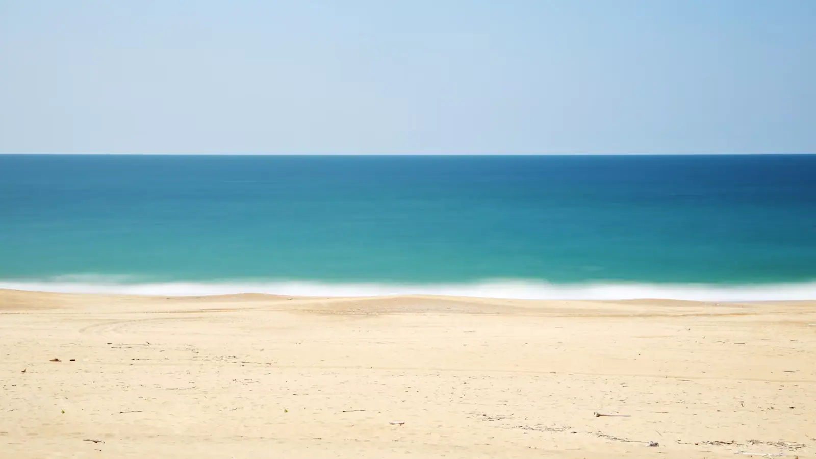 An empty stretch of pale sand meeting calm shallow water under a clear sky