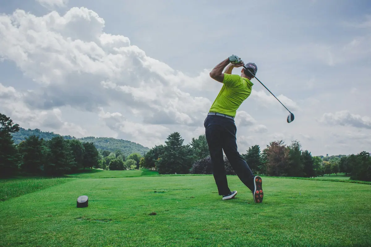 A golfer finishing a drive from an elevated tee on a wooded course under a cloudy sky