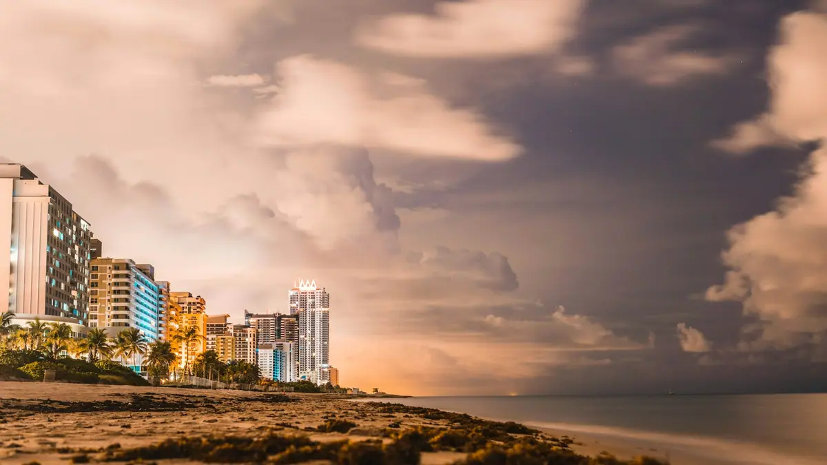 A storm cloud building over a Gulf beach at dusk with lit condominium towers along the shoreline