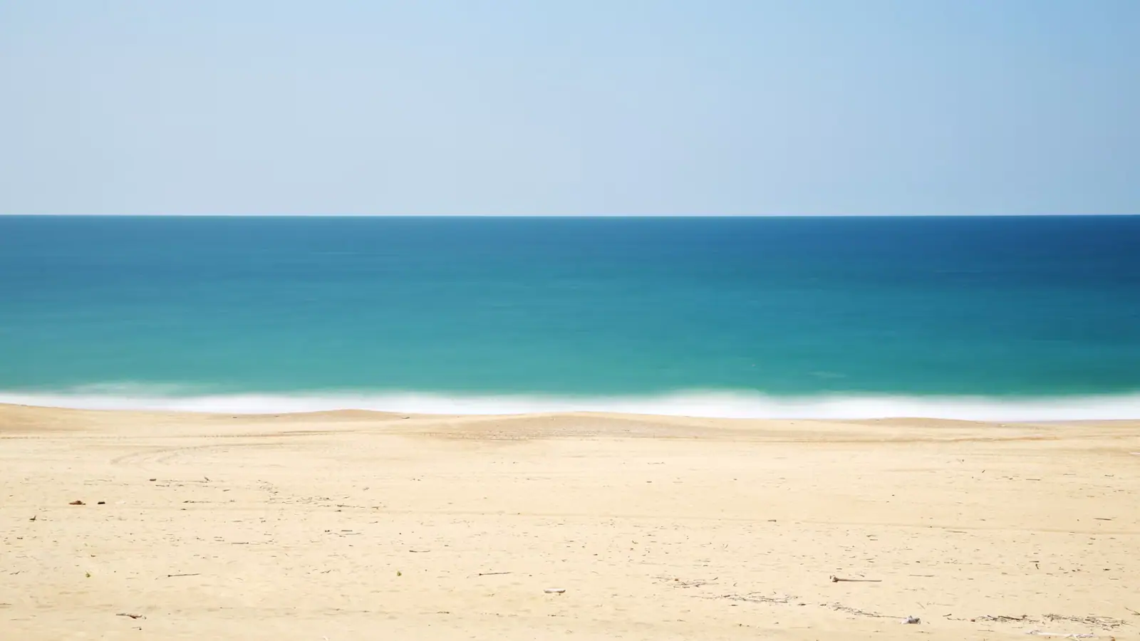 An empty stretch of pale sand meeting calm shallow water under a clear sky