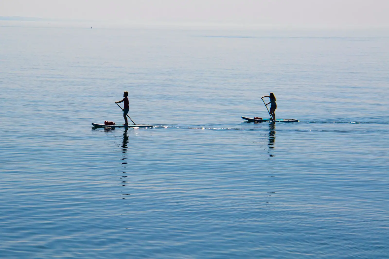 Two paddleboarders crossing a wide sheet of flat, mirror-calm water under a soft sky