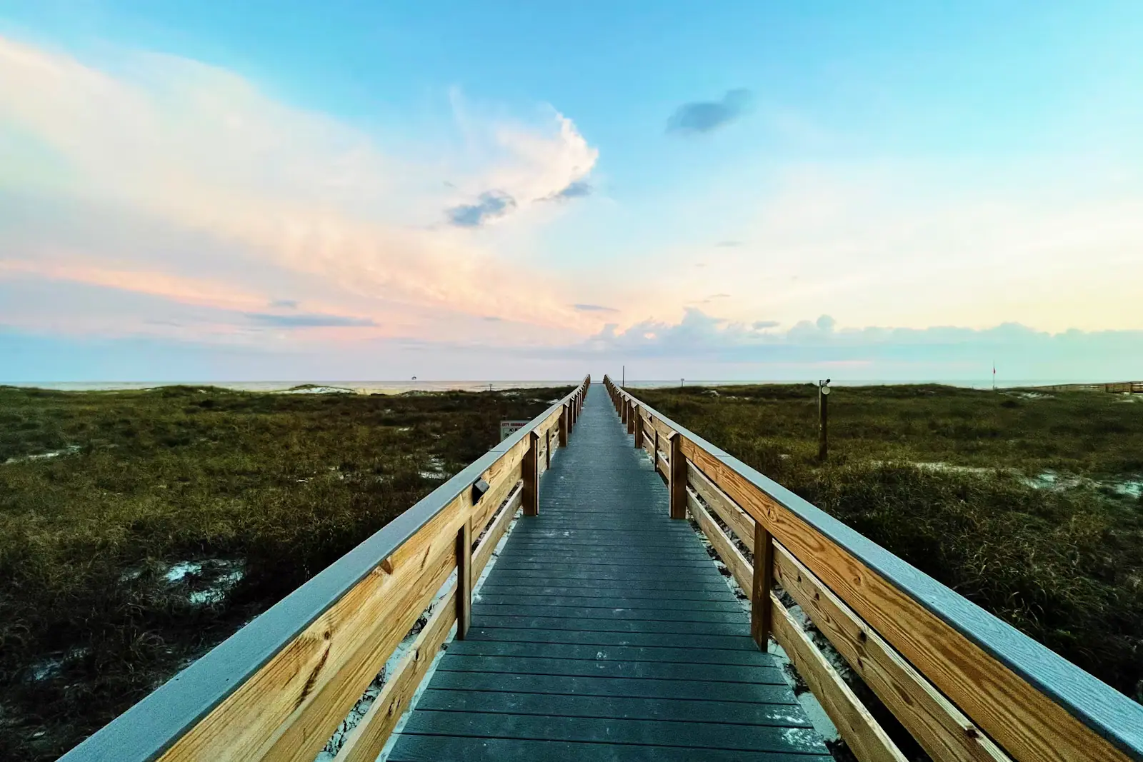 A wooden boardwalk crossing pale beach sand towards the water under an evening sky