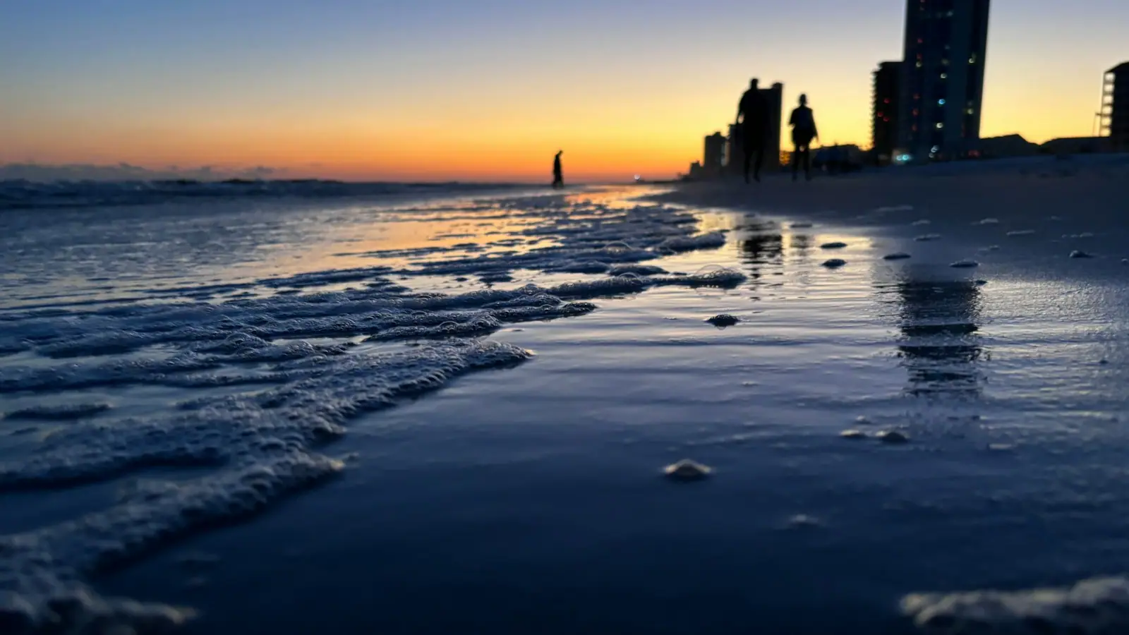 A beach at sunset with low buildings silhouetted along the shoreline behind it
